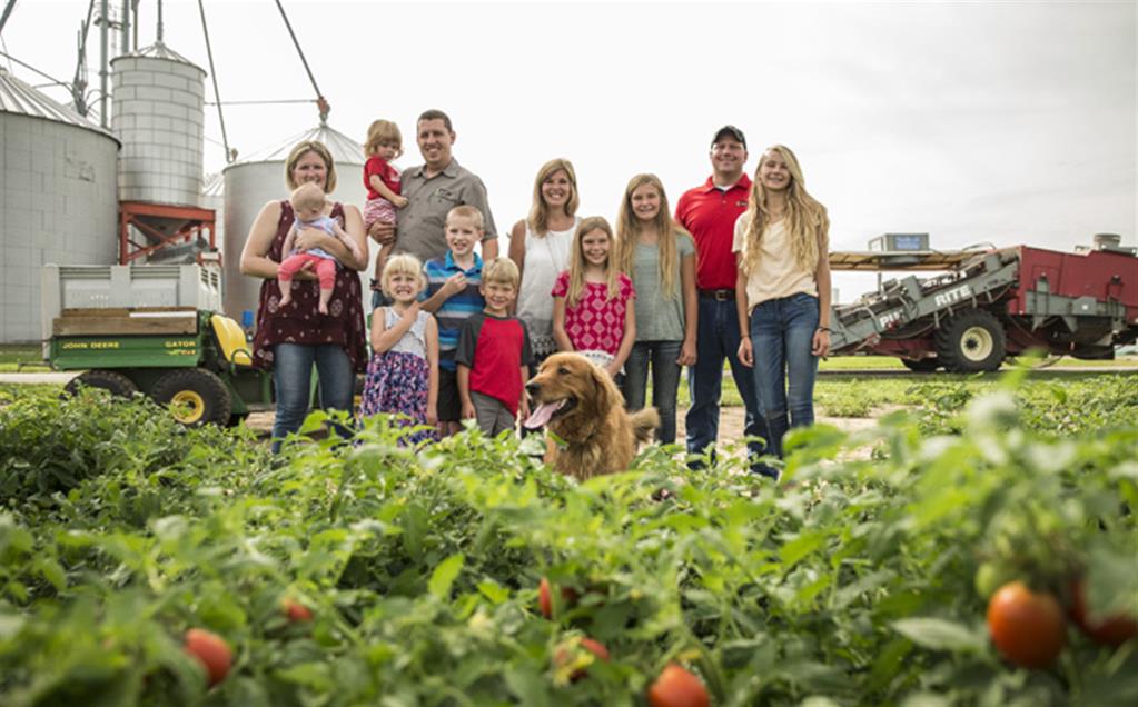 Image of Red Gold Growers Eck Family Farms from Boggstown Indiana families standing by tomato field with harvester in background and grain silos