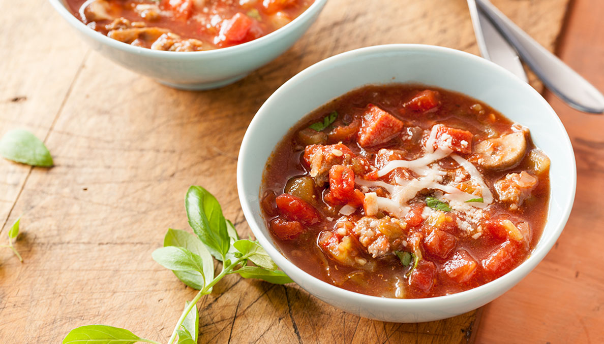 Image of a bowl of Cheesy Pizza Soup with italian sausage, canned diced tomatoes, sliced mushrooms and cheese in a white bowl on a wood surface