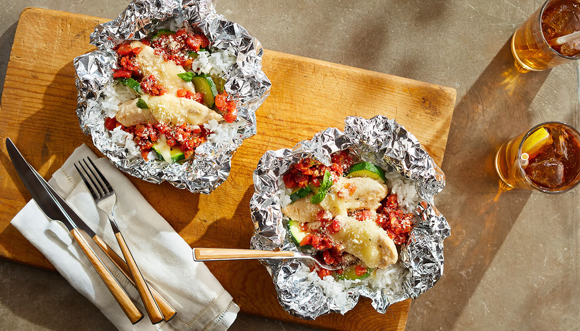 Overhead image of Italian Chicken Foil Packet Recipe in tin foil on a wood cutting board with iced tea and silverware on the side Red Gold tomatoes