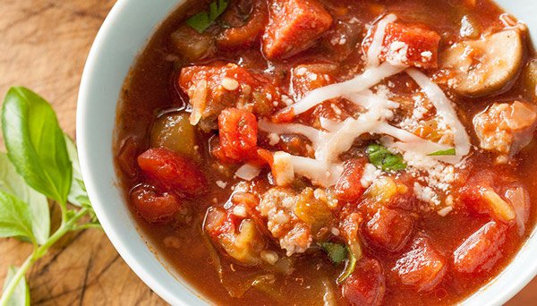 Overhead image of pizza soup with canned diced tomatoes sliced mushrooms italian sausage pepperoni and cheese in a white bowl next to fresh basil